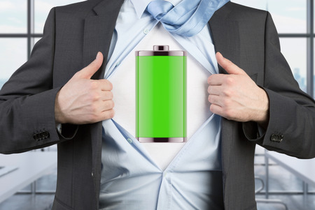 A man in a suit is tearing the blue shirt. Full charged battery on the chest. Modern panoramic office background.の写真素材