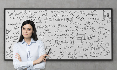 A beautiful brunette is pondering about the solution of complicated analytical problem. Math formulas are written down on the whiteboard. Concrete wall.の写真素材