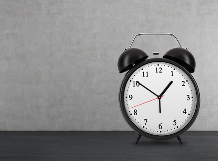 A black alarm clock is on the black floor. Concrete wall.