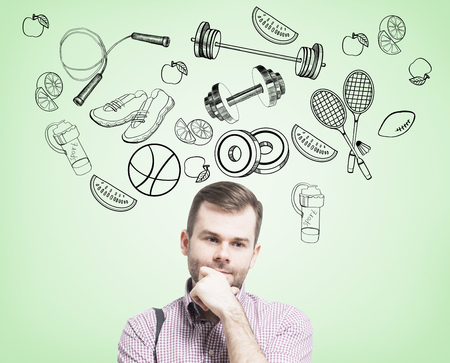Pondering handsome man in casual clothes is thinking about his choice of sport activity. Sport icons are drawn on the concrete wall.の写真素材