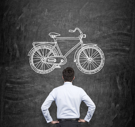 Rear view of a businessman in formal clothes who is looking at the huge black chalkboard with a drawn sketch of a bicycle. A concept of environmental friendly ways of commuting and travelling.の写真素材