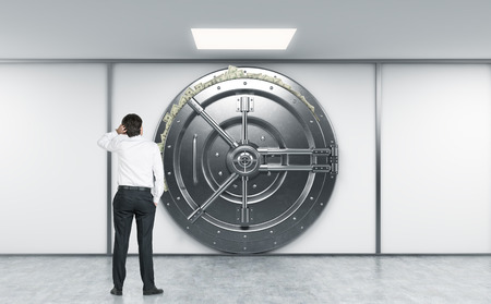 man sratching head with left hand standing in front of a big locked round metal safe in bank depository with dollars stuck out from behind the door, concept of abundance,  front view with man's backの写真素材