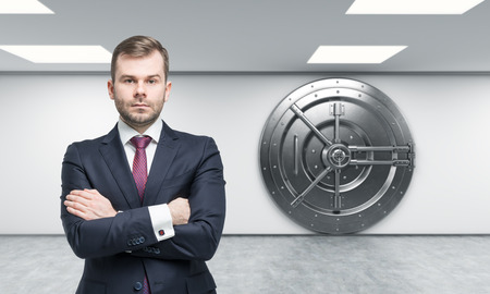 businessman with arms crossed standing in front of a big locked round metal safe in a bank depository,  a concept of security,  front view,の写真素材