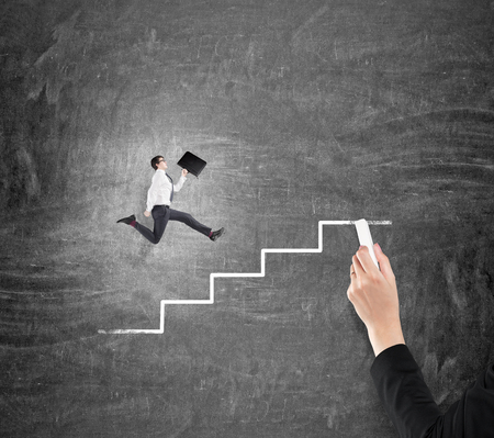 Young businessman with a folder running up a drawn stairs along a blackboard, hand drawing steps with a chalk. Concept of career growth.の写真素材