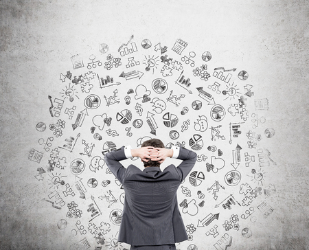 Businessman with his hands on the back of his head looking at the concrete wall with small business icons. Concept of work.の写真素材