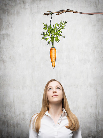 A young pretty woman looking upwards at a painted carrot tied to a branch. Concrete background. Front view. Concept of reward.の写真素材