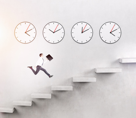 A young man with a folder running upstairs, four clocks on the white wall over seven steps. Concept of career growth.の写真素材