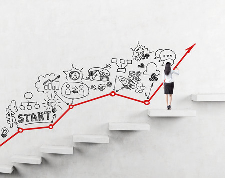 woman on a step of a ladder painting black icons describing business development on the white wall. Red graph on the wall. Back view. Concept of business development.の写真素材