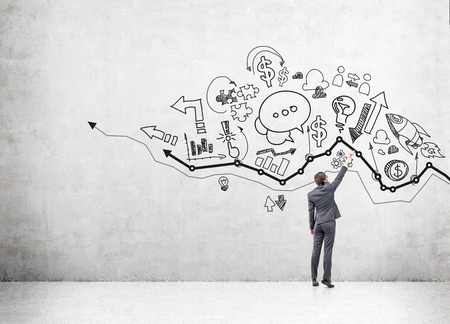 A young businessman drawing many different business icons on a concrete wall over a graph. Back view. Concept of business development.の写真素材