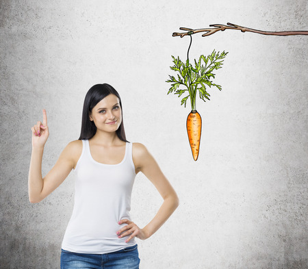 A young pretty woman with an index finger up standing next to a picture of a carrot tied to a branch. Concrete background. Concept of a new idea and future praise.の写真素材