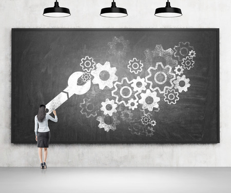 A businesswoman drawing a wrench and numerous cog wheels on a blackboard. Three black lamps over it. Back view. Concrete background. Concept of fixing things.の写真素材