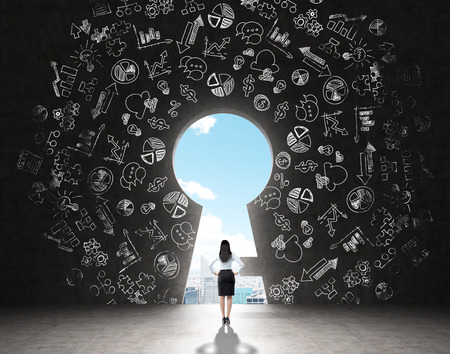 A woman with hands on hips standing in front of a huge keyhole, many business icons drawn around it, city and sky seen through it. Black background. Back view.  Concept of finding the wayの写真素材