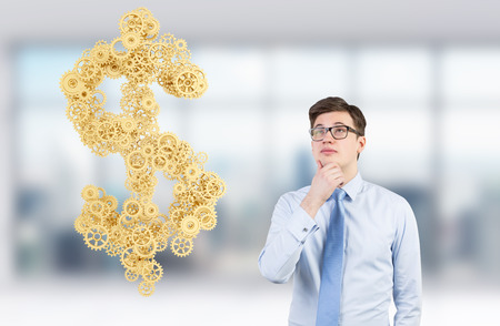 A young businessman with hand to the chin imagining a huge dollar symbol composed of many golden cogwheels. Blurred office background. Concept of getting money.の写真素材
