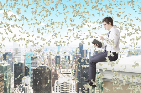 A young man sitting on the edge of the roof and reading a book, money falling from above. Side view. New York at the background. Concept of studyingの写真素材