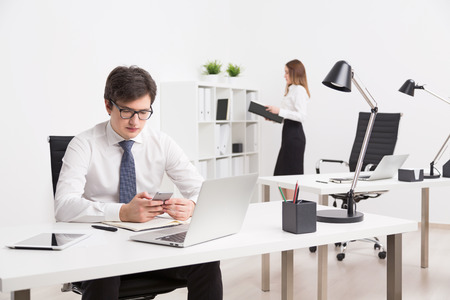 Businessman with smartphone in front of laptop, businesswoman at background. Office. Concept of work.の写真素材