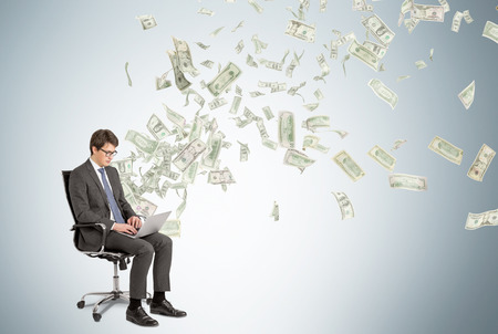 Businessman with laptop sitting on chair, dollars flying from behind. Grey background. Concept of making money.の写真素材