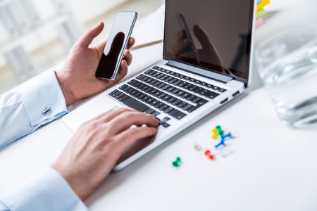 Businessman sitting at computer, smartphone in hand, office at background. Concept of work.の写真素材