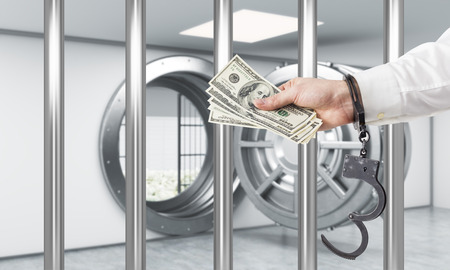 Hand in handcuffs holding dollars in front of open safe door behind bars. Concept of stealing money.の写真素材