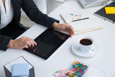 Businesswoman sitting at table. Tablet, notebook and cup in front of her. Only hands seen. Office. Concept of work.の写真素材