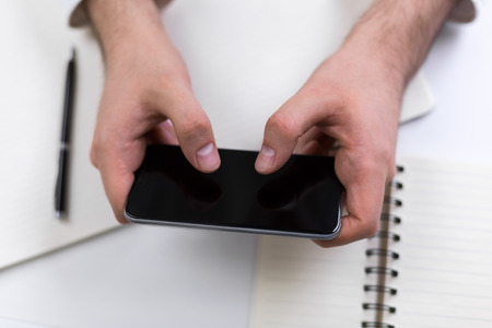 Closeup of businessman hands using blank smartphone above desktop with office tools. Mock up