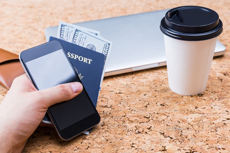 Cork table with closed laptop, coffee cup and hand holding smartphone and american passport with dollars. Traveling conceptの写真素材