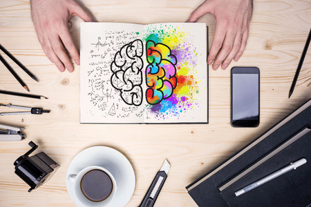 Top view of wooden desktop with coffee cup, stationery items, smart phone and hands holding notepad with creative and analytical brain sides sketchの写真素材