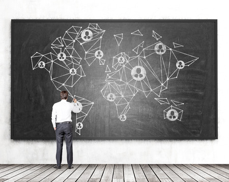 Concept of global social network with businessman drawing abstract polygonal map and people icons on chalkboard in room with concrete wall and wooden floorの写真素材