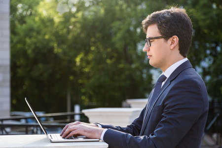Side view of handsome young businessman using laptop outside on blurry green tree backgroundの写真素材