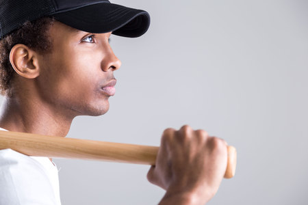 Closeup of attractive african american guy in cap holding baseball bat and looking away from the camera on light backgroundの写真素材
