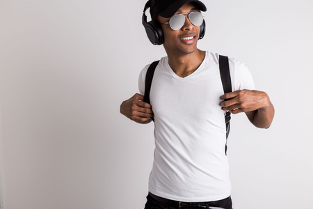 Handsome cheerful african american guy in cap and sunglasses listening to music with headphones on light backgroundの写真素材