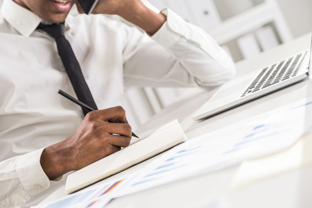 Side view of attractive african american businessman using notepad and notebook on office desktop while having mobile phone conversation. Bookshelf with documents in the backgroundの写真素材
