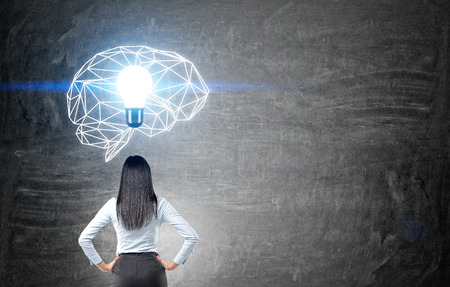Brainstorming concept with thoughtful businesswoman looking at chalkboard with illuminated polygonal brain sketch and abstract light bulbの写真素材