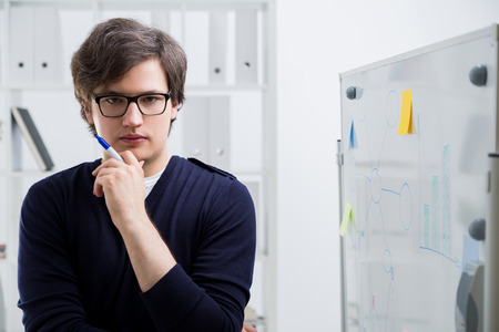 Concentrated young man standing next to office whiteboard with business charts and looking at the...