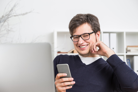 Attractive smiling businessman in glasses using cellphone at his workplaceの写真素材