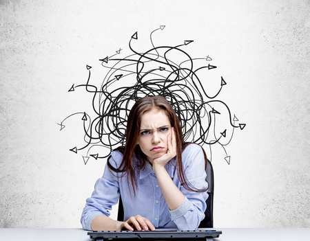 Grumpy young girl using keyboard against concrete wall with tangled arrows sketch. Different direction conceptの写真素材