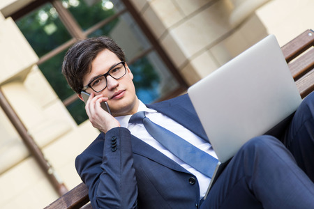 Handsome young businessman in suit sitting on wooden bench with notebook placed on his laptop, talking on the phoneの写真素材