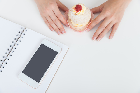 Woman hand holding cupcake. Her smart phone lies on top of notebook on table. Concept of tasty cupcake eating during lunch break and difficulties of dieting. Close up. Top viewの写真素材