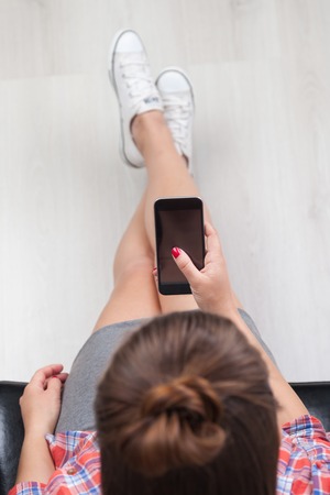 Girl sitting with her legs crossed in leather armchair looking at smart phone screen. Concept of communication. Top viewの写真素材