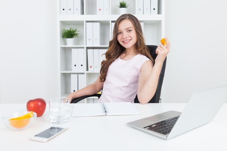Woman holding orange and smiling. She is at her working place in company office. Big red apple and slice of orange lie on desk near glass of water, cell phone and laptop. Concept of good workspace.の写真素材