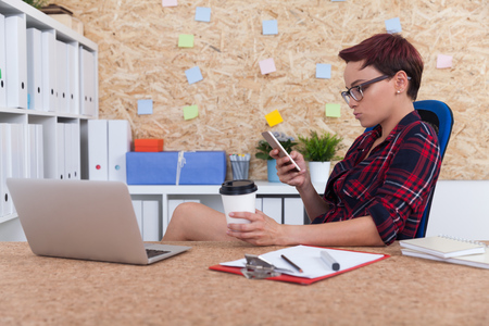 Office employee checking her phone during lunch hour and drinking coffee. Concept of work and rest in companyの写真素材