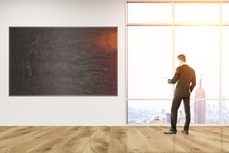 Man in suit standing in office corridor with blackboard on wall looking at city view through big window. Concept of management. 3d rendering. Mock up. Toned image.の写真素材