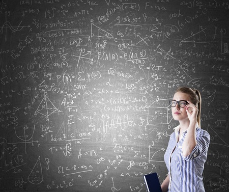 Nerd girl standing near blackboard with formulas and figures. Concept of engineering and exact sciences.の写真素材