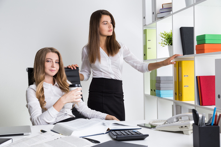 Blond business lady is enjoying her coffee. Her colleague is taking large yellow binder from shelf. Concept of clerk jobの写真素材