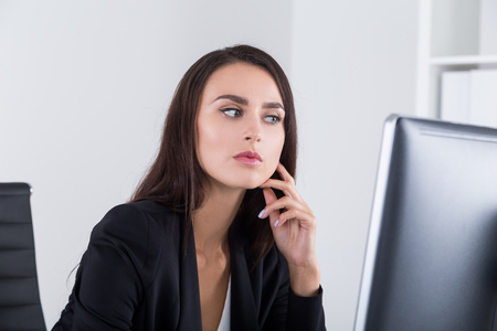 Pensive business lady is sitting at her workplace near large computer monitor and looking at it analyzing data. Concept of information analysis in business.の写真素材