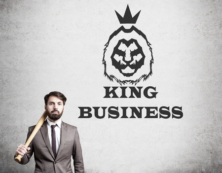 Businessman with baseball bat is standing near concrete wall with king business text on it. Concept of high league businessmanの写真素材