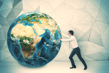 Side view of businessman in glasses pushing the globe in room with concrete walls and geometric ornament. Concept of significant project. Toned image.の写真素材