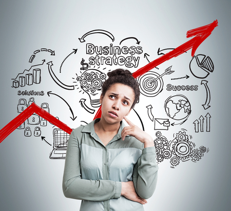 Portrait of confused African American businesswoman standing against gray wall with red graph and startup sketch on it. Concept of female small business owner.の写真素材