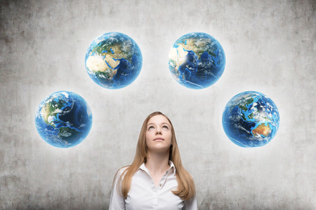 Blond girl standing against concrete wall background and looking at four globes showing different parts of the world. Concept of globalization and international business companiesの写真素材