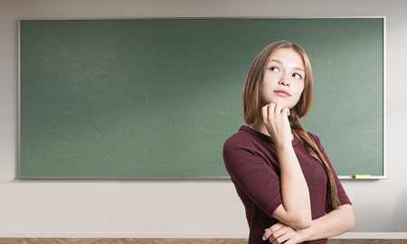 Portrait of young girl with braided hair standing in class with empty green chalkboard. Concept of education and opportunities. Mock upの写真素材