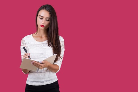 Serious girl in white blouse standing against red background and writing in her notebook with spring. Concept of journalism. Mock upの写真素材